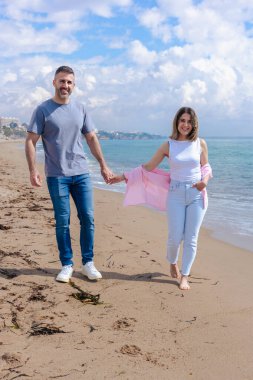 Kumsalda çıplak ayakla yürüyen, el ele tutuşan ve tatilde gülümseyen beyaz bir çift. Okyanus dalgaları, kum ve arka planda parlak bir gökyüzü olan sakin bir deniz sahili anı.