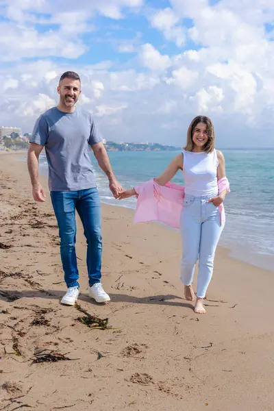 Kumsalda çıplak ayakla yürüyen, el ele tutuşan ve tatilde gülümseyen beyaz bir çift. Okyanus dalgaları, kum ve arka planda parlak bir gökyüzü olan sakin bir deniz sahili anı.