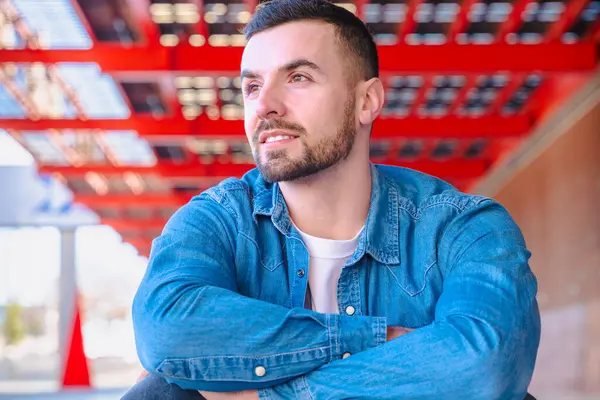 Modern bir şehir ortamında düşünceli bir şekilde oturan beyaz bir gencin portresi. Sıradan bir kot giysisi, yukarıda kırmızı mimari elementler ve doğal ışık şık ve rahat bir şehir havası yaratır.. 