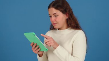 Beyaz bir kadın, elinde yeşil bir tablet tutarken ve gülümserken görülmüş. Günlük beyaz süveter, mavi arka plan ve temiz fotokopi alanı burayı modern ve minimal teknoloji sahnesi yapar. 