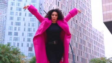 Pembe palto giyen kıvırcık saçlı Afrikalı Amerikalı dansçı şehir ortamında hareketlerini ve enerjilerini ifade ediyor. Modern moda, çağdaş dans ve sanatsal performans şehrin arka planına karşı.