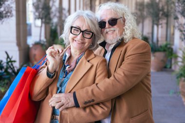Kameraya gülümseyen yaşlı beyaz bir çift, canlı alışveriş torbaları tutuyor ve sarılıyorlar. Şık paltolarını giyerek şık bir açık hava alışveriş alanında keyifli bir anın tadını çıkarırlar.. 