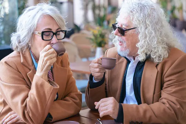 Gözlüklü ve kahverengi paltolu yaşlı bir çift açık hava kafesinde kahvenin tadını çıkarıyor. Sıcak bir anı paylaşıyorlar, gülümsüyorlar ve gündüzleri huzurlu, rahat bir atmosferde rahatlıyorlar.. 
