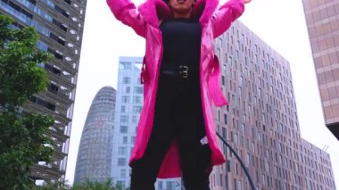 Pembe palto giyen kıvırcık saçlı Afrikalı Amerikalı dansçı şehir ortamında hareketlerini ve enerjilerini ifade ediyor. Modern moda, çağdaş dans ve sanatsal performans şehrin arka planına karşı.