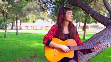 Uzun saçlı genç bir kadın güneşli bir parkta akustik gitar çalıyor, bir ağacın altında huzur içinde oturuyor. Doğal ortam, rahat atmosfer, müzik ve boş zaman atmosferi.