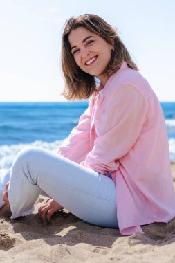 Kumlu bir plajda oturmuş, arkasında okyanus varken kameraya gülümseyen mutlu genç bir kadının dikey portresi. Rahat bir ortam, günlük kıyafetler, doğal ışık ve sakin bir ortam.. 