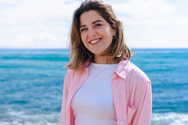 Deniz kenarında pembe bir gömlek giymiş gülümseyen genç bir kadının yatay portresi. Arka planda berrak mavi okyanus, günlük tarz, doğal ışık ve dışarıdaki kameraya bakan neşeli ifade.. 