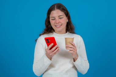 Günlük kıyafetli genç beyaz kadın gülümserken bir kahve fincanı ve akıllı telefon tutuyor. Mutlu ve heyecanlı görünüyor. Mavi bir zemine karşı duruyor. Neşeli ve rahat bir anın tadını çıkarıyor..