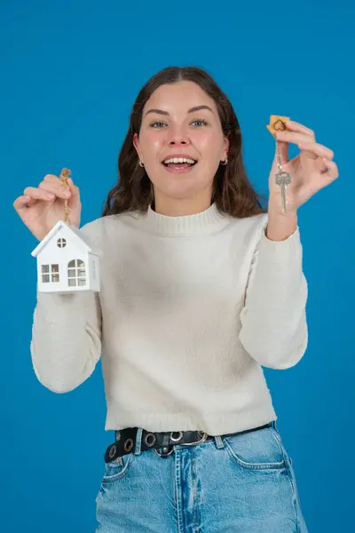 Evin modelini ve anahtarlarını tutan mutlu genç bir kadın heyecanını dile getiriyor. Beyaz bir kazak giyiyor ve mavi bir arka plana karşı duruyor. Ev sahipliği, gayrimenkul veya yatırım kavramı. dikey