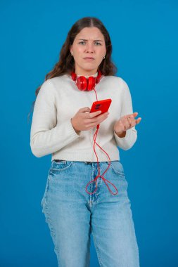 Kafkasyalı bir kadının akıllı telefonuyla video izlerken çekilmiş dikey portresi. Kulaklık takıyor ve günlük giysiler giyiyor, temiz mavi bir arka plana karşı dijital bir anın tadını çıkarıyor.. 