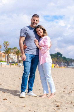 Tatil sırasında sahilde kucaklaşan gülümseyen beyaz bir çiftin dikey portresi. Deniz kenarında sakin ve sevecen bir an. Mavi gökyüzü, okyanus dalgaları ve arkalarında kumlu bir sahil.. 