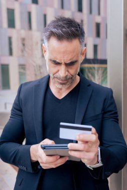 Kafkasyalı yetişkin bir adam tablet kullanıyor ve elinde online ödeme yapmak için bir kredi kartı tutuyor. Modern finans, alışveriş ve dijital yaşam tarzını temsil eden kentsel bir ortamda odaklanmış ifade. Dikey