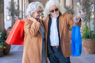 Neşeli son sınıf çifti alışveriş yaptıktan sonra renkli çantaları havada tutarak kutlama yapıyor. Şık paltolar giyip, dışarıda bitkilerle dolu büyüleyici bir şehir ortamında dururken mutlu bir şekilde gülümsüyorlar..