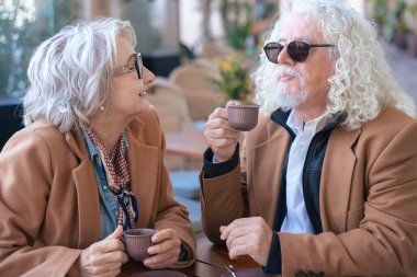 Gözlüklü ve kahverengi paltolu yaşlı bir çift açık hava kafesinde kahvenin tadını çıkarıyor. Sıcak bir anı paylaşıyorlar, gülümsüyorlar ve gündüzleri huzurlu, rahat bir atmosferde rahatlıyorlar..