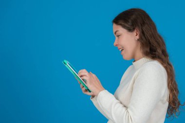 Beyaz kazak giyen gülümseyen genç bir kadının yeşil tablet kullanarak fotokopi alanındaki mavi arka plana bakması dijital iletişimi, taramayı ve teknolojik etkileşimi sembolize ediyor.. 