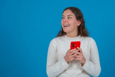 Beyaz kazaklı, gülümseyen ve elinde kırmızı bir akıllı telefon tutan genç bir kadın, soluna bakarken, canlı mavi bir arka planda duruyor, merakı, teknolojiyi ve olumlu duyguyu temsil ediyor.. 