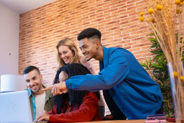 Çok kültürlü bir grup genç arkadaş bir dizüstü bilgisayarın etrafında toplandı. Sıcak, kapalı bir ortamda, tuğla duvar ve bitkilerle gülümsüyor ve etkileşime giriyorlardı. Çeşitliliği, dostluğu ve bağlantıyı temsil ediyorlardı.. 