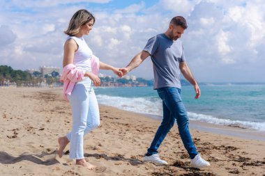 Kumsalda çıplak ayakla yürüyen, el ele tutuşan ve tatilde gülümseyen beyaz bir çift. Okyanus dalgaları, kum ve arka planda parlak bir gökyüzü olan sakin deniz kenarı anı. Boşluğu kopyala. 