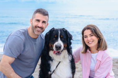 Tatil sırasında kumsalda büyük bir köpekle gülümseyen beyaz bir çiftin portresi. Deniz kenarında rahat ve keyifli bir an. Üçü de aydınlık bir günde kameraya bakıyor.. 