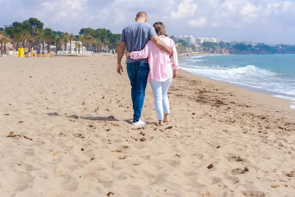 Mavi gökyüzünün altında kumlu bir sahil boyunca kol kola yürüyen sevgi dolu bir çiftin arka görüntüsü sahil ortamında romantizmi, bağlantıyı, barışı ve rahatlamayı simgeliyor.. 