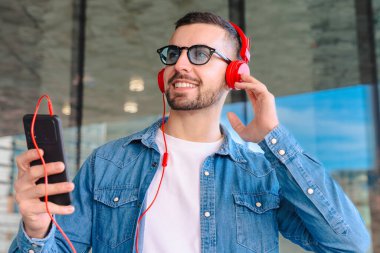 Genç adam, güneş gözlüğü ve kırmızı kulaklık takıyor. Modern cam bir ortamda dışarıda dururken akıllı telefonundan müziğin keyfini çıkarıyor. Yaşam tarzını, teknolojisini ve rahatlamasını ses yoluyla temsil ediyor.. 