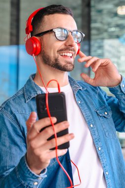 Genç adam, güneş gözlüğü ve kırmızı kulaklık takıyor. Modern cam bir ortamda dışarıda dururken akıllı telefonundan müziğin keyfini çıkarıyor. Yaşam tarzını, teknolojisini ve rahatlamasını ses yoluyla temsil ediyor.. 