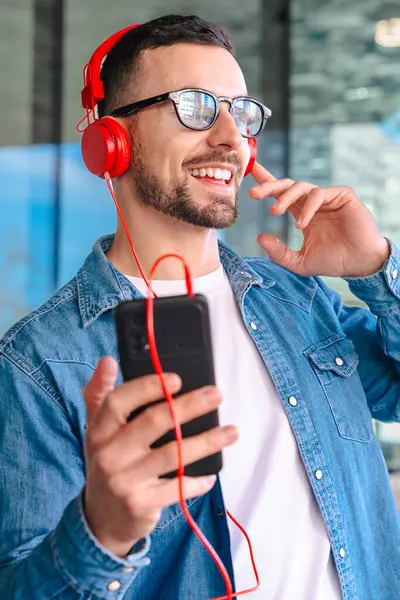 Genç adam, güneş gözlüğü ve kırmızı kulaklık takıyor. Modern cam bir ortamda dışarıda dururken akıllı telefonundan müziğin keyfini çıkarıyor. Yaşam tarzını, teknolojisini ve rahatlamasını ses yoluyla temsil ediyor.. 