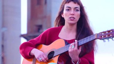 Genç bir kadın, akustik gitar çalıp şarkı söylerken, kırmızı bir kazak giyerken ve duygularını ifade ederken, canlı müzik, yaratıcılık, tutku ve doğal sanatsal ifadeyi temsil ederken, dışarıda sahneye çıkıyor.. 