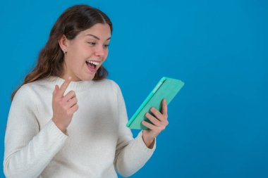 Beyaz kazaklı heyecanlı genç bir kadın, yeşil bir tablete bakarken şaşkınlıkla tepki veriyor, tek parmakla işaret ediyor, fotokopi çekilen mavi arka planda, keşfi ya da haberleri simgeliyor.. 