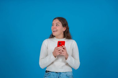 Beyaz kazaklı, gülümseyen ve elinde kırmızı bir akıllı telefon tutan genç bir kadın, soluna bakarken, canlı mavi bir arka planda duruyor, merakı, teknolojiyi ve olumlu duyguyu temsil ediyor.. 