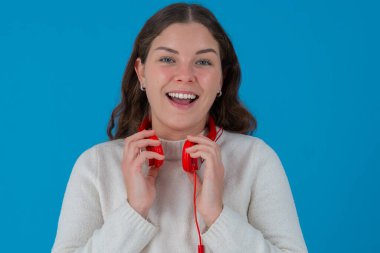 Boynuna kırmızı kulaklık takmış, mutlu bir şekilde gülümseyen ve rahatlamış görünen bir kadın. Neşeli ve kendinden emin görünüyor. Düz mavi arkaplan. 