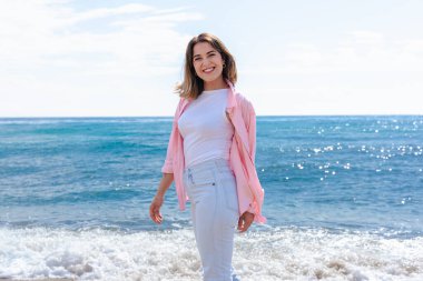 Kumsalda durup gülümseyerek kameraya bakan mutlu genç bir kadın. Günlük giysiler içinde güneşli bir günün ve huzurlu bir kıyı manzarasının keyfini çıkarıyor.. 