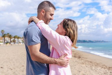 Romantik Kafkasyalı çift sahilde kucaklaşırken birlikte vakit geçiriyorlar. Parlak bir gökyüzü altında birbirlerine sevgiyle gülümserler. Etrafları kum, deniz ve rahat bir yaz atmosferiyle çevrilidir.. 