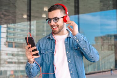 Genç adam, güneş gözlüğü ve kırmızı kulaklık takıyor. Modern cam bir ortamda dışarıda dururken akıllı telefonundan müziğin keyfini çıkarıyor. Yaşam tarzını, teknolojisini ve rahatlamasını ses yoluyla temsil ediyor.. 