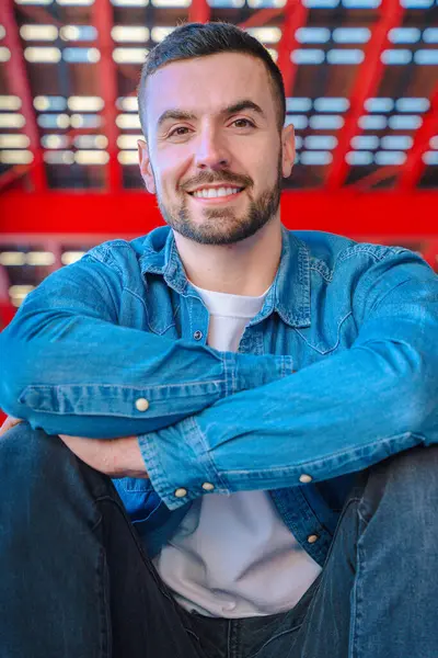 Kendinden emin beyaz bir adam, sıradan bir şekilde oturuyor ve dikey bir portrede kameraya gülümsüyor. Kot ceket giyiyor ve rahat bir tavırla modern kırmızı şehir geçmişine karşı poz veriyor.. 