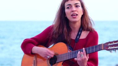 Akustik gitar çalan ve deniz kenarında şarkı söyleyen sanatçı genç bir kadın. Doğayla ve yumuşak doğal ışıkla çevrili sakin bir sahil ortamında duygusal bir performans sergiliyor.. 