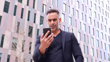 Hayal kırıklığına uğramış bir adam akıllı telefon ekranını şehir ortamında doğrudan kameraya doğrultuyor. Öfkeli ifadesi cihaz, uygulama ya da dijital iletişimde bir sorun olduğunu gösteriyor.. 