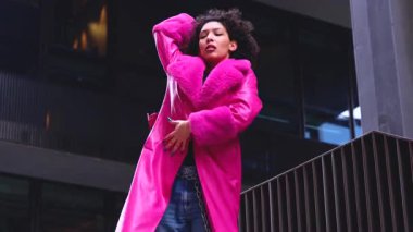 Pembe palto giyen kıvırcık saçlı Afrikalı Amerikalı dansçı şehir ortamında hareketlerini ve enerjilerini ifade ediyor. Modern moda, modern dans ve sanatsal performans.