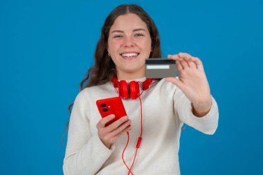 Beyaz kazaklı ve kırmızı kulaklıklı mutlu genç bir kadın kameraya bir kredi kartı gösterirken gülümsüyor ve elinde kırmızı bir akıllı telefon tutuyor, mavi arka planda online alışverişi sembolize ediyor.. 