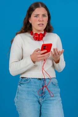 Kafkasyalı bir kadının akıllı telefonuyla video izlerken çekilmiş dikey portresi. Kulaklık takıyor ve günlük giysiler giyiyor, temiz mavi bir arka plana karşı dijital bir anın tadını çıkarıyor.. 