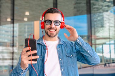 Genç adam, güneş gözlüğü ve kırmızı kulaklık takıyor. Modern cam bir ortamda dışarıda dururken akıllı telefonundan müziğin keyfini çıkarıyor. Yaşam tarzını, teknolojisini ve rahatlamasını ses yoluyla temsil ediyor.. 