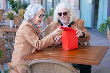 Gözlüklü ve kahverengi paltolu yaşlı bir çift açık hava kafesinde kahvenin tadını çıkarıyor. Sıcak bir anı paylaşıyorlar, gün içinde huzurlu ve rahat bir atmosferde gülümsüyorlar. Dikey