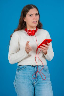 Kafkasyalı bir kadının akıllı telefonuyla video izlerken çekilmiş dikey portresi. Kulaklık takıyor ve günlük giysiler giyiyor, temiz mavi bir arka plana karşı dijital bir anın tadını çıkarıyor.. 