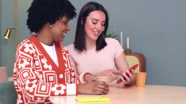 Neşeli Kafkasyalı ve Afro-Amerikalı kadınlar koltukta oturmuş kırmızı bir akıllı telefona bakarken eğleniyorlar, kahkahaları paylaşıyorlar ve içeride kaliteli zaman geçiriyorlar. 