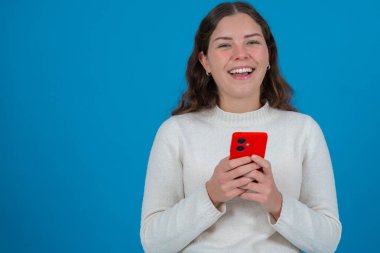 Beyaz kazaklı, kırmızı bir akıllı telefon tutan ve doğrudan kameraya gülümseyen mavi arka planda duran, iletişimi ve güveni sembolize eden neşeli genç bir kadın portresi. Boşluğu kopyala