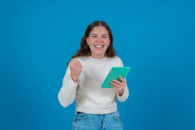 Beyaz kazaklı mutlu genç bir kadın yeşil bir tablet tutarken yumruğunu kaldırıyor ve kameraya gülümsüyor. Mavi arka plan, başarıyı, neşeyi ve dijital hayatı simgeliyor.. 