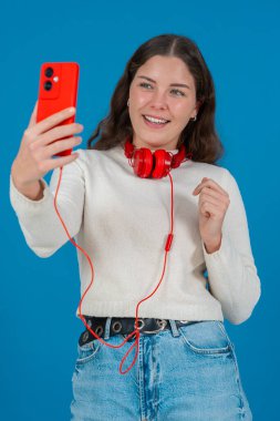 Kırmızı akıllı telefon ve kulaklıkla selfie çeken mutlu genç bir kadının dikey görüntüsü. Mavi bir arkaplan önünde, gündelik ve modern bir dijital anın tadını çıkararak güvenle gülümsüyor. Dikey