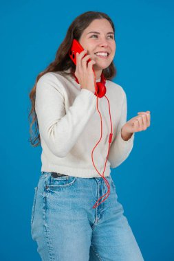 Kırmızı bir akıllı telefondan konuşan neşeli genç bir kadının dikey görüntüsü. Elini göğsüne koyup gülüyor. Parlak mavi bir arka plana yaslanıyor. Sıradan bir anda.. 