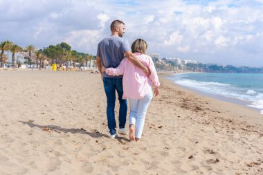 Mavi gökyüzünün altında kumlu bir sahil boyunca kol kola yürüyen sevgi dolu bir çiftin arka görüntüsü sahil ortamında romantizmi, bağlantıyı, barışı ve rahatlamayı simgeliyor.. 