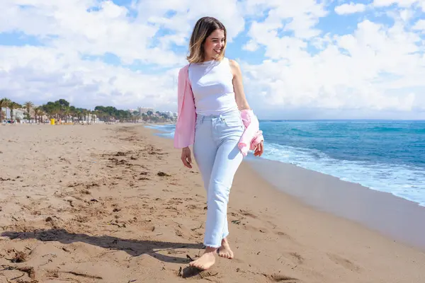 Gülümseyen genç bir kadın güneşli bir günde kumlu bir sahilde çıplak ayakla yürür. Hafif kıyafetler giyer ve etrafındaki dalgalar, mavi gökyüzü ve doğal güzelliklerle huzurlu kıyı atmosferini sever.. 